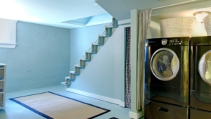 Basement laundry area with stacked washer dryer, stairs, light blue walls, and small rug