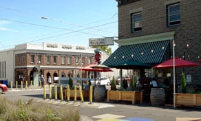 Outdoor café with striped awning, red umbrellas, and string lights on a sunny street corner.