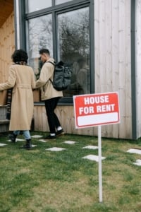 Young couple carrying boxes towards a modern rental home with a House for Rent sign in a residential neighborhood.